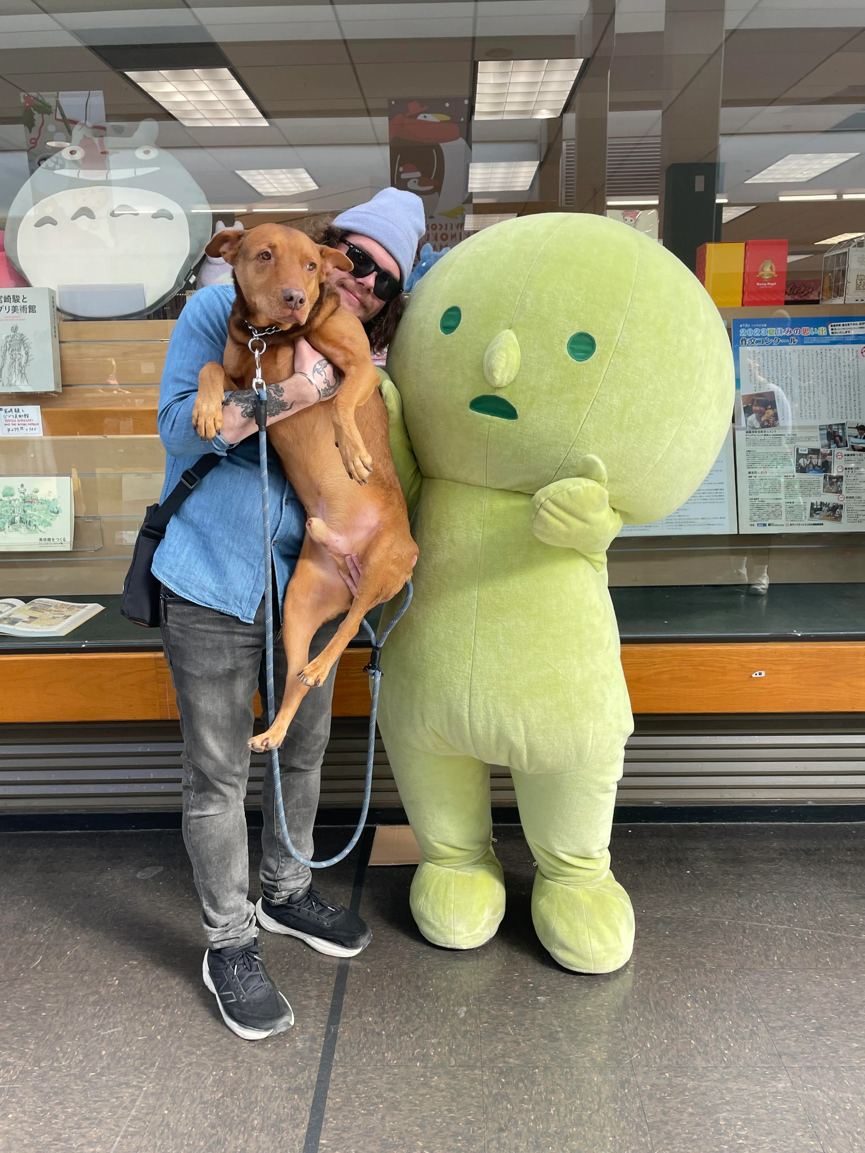 Jacob with a brown dog, hugging a life-size green Smiski mascot in front of a shop window.